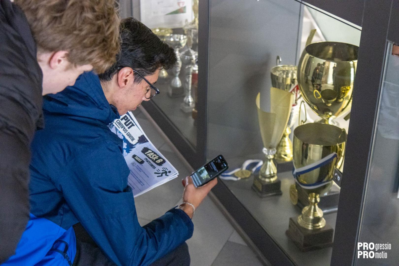 Two students look at trophies in a display case and take a picture with a smartphone, holding a task sheet titled "PUT CAMPUS GAME TASK B".