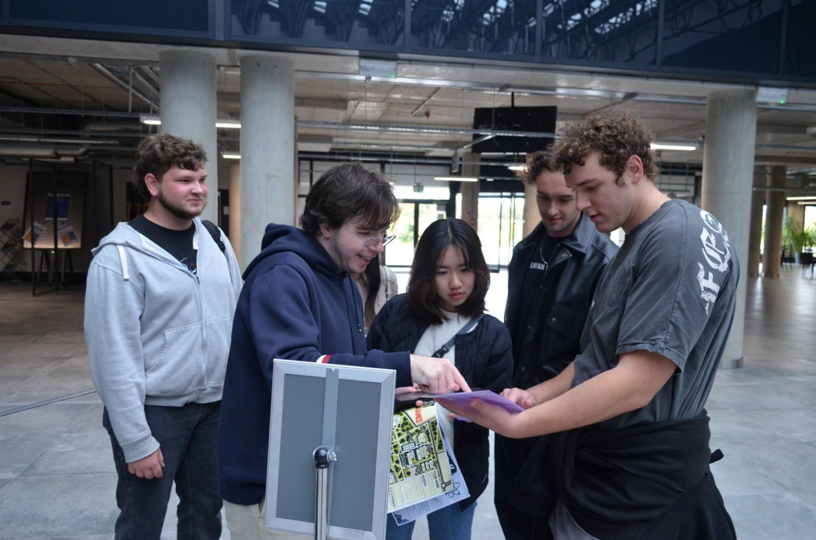 A group of students in the spacious hall of the building analyzes a purple task sheet and the event map of the PUT Campus Game.
