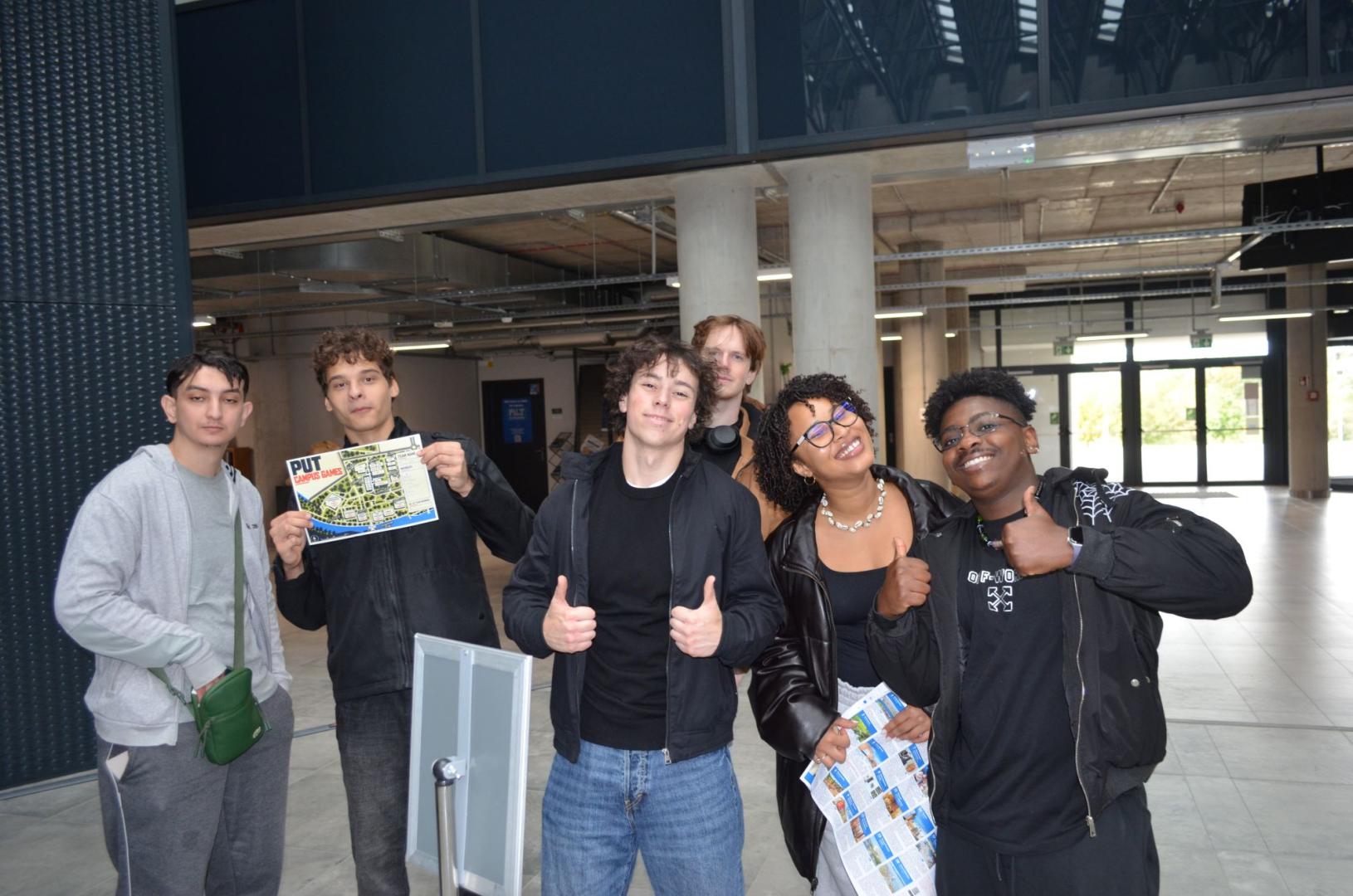 A smiling group of international students poses in a modern hall, giving thumbs up and holding a "PUT Campus Game" map.