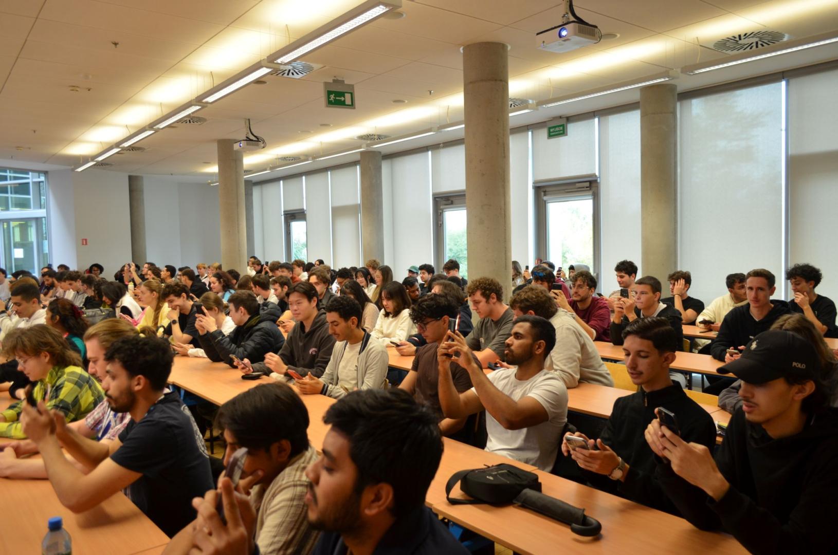 An audience full of students sitting at desks in a large lecture hall uses their smartphones.