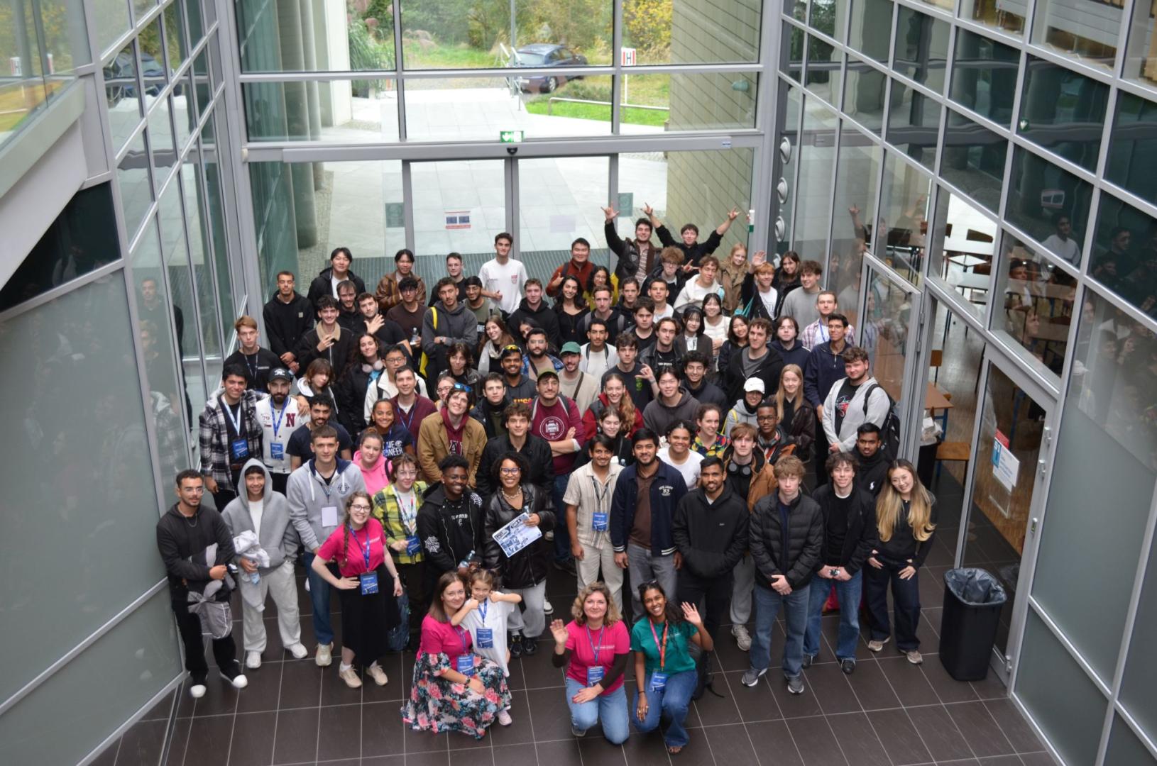 A large group of international students and organizers poses for a commemorative photo in the glass-walled hall of the university building.
