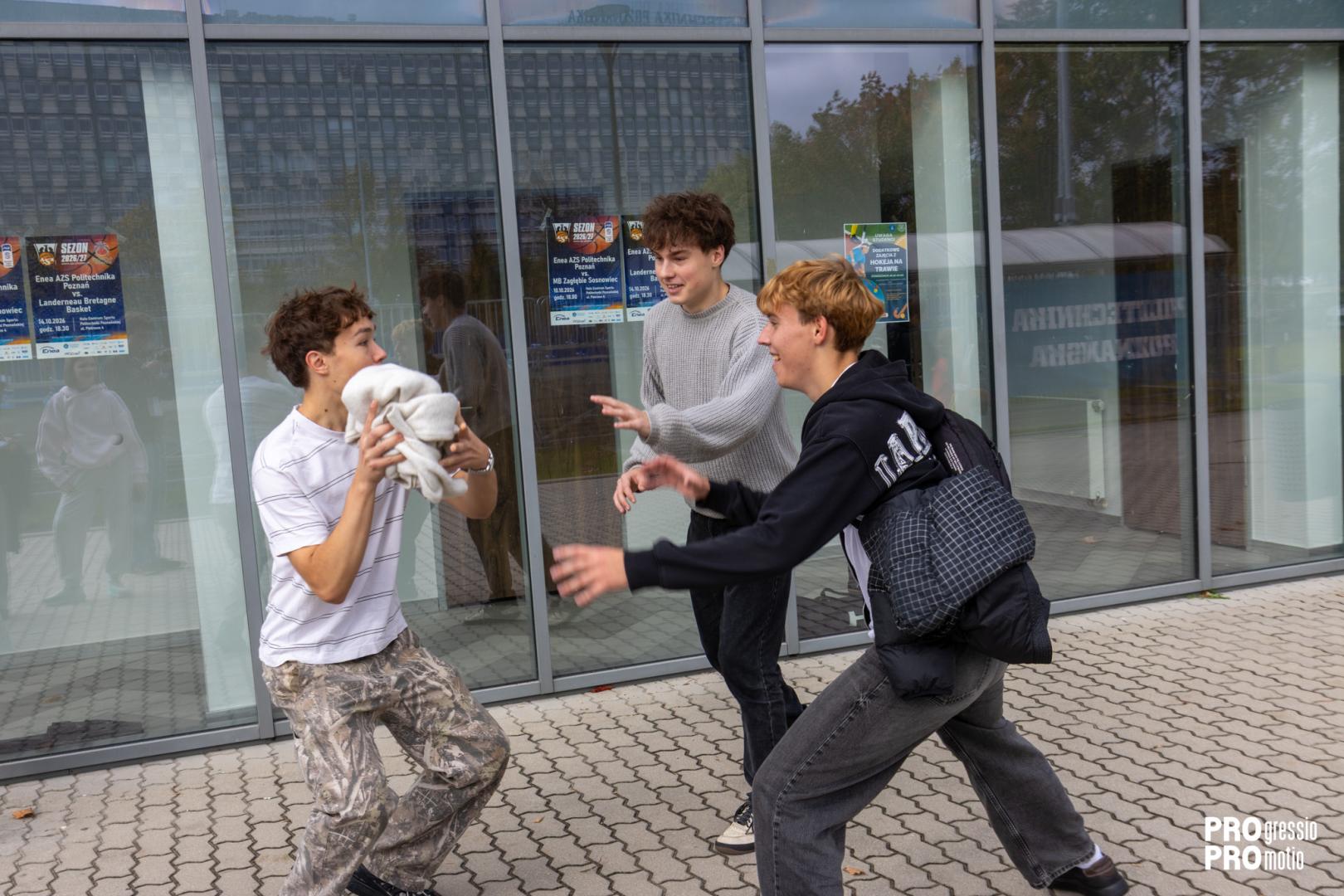 Three young students play dynamically in front of the glass facade of the university building, performing one of the campus game tasks