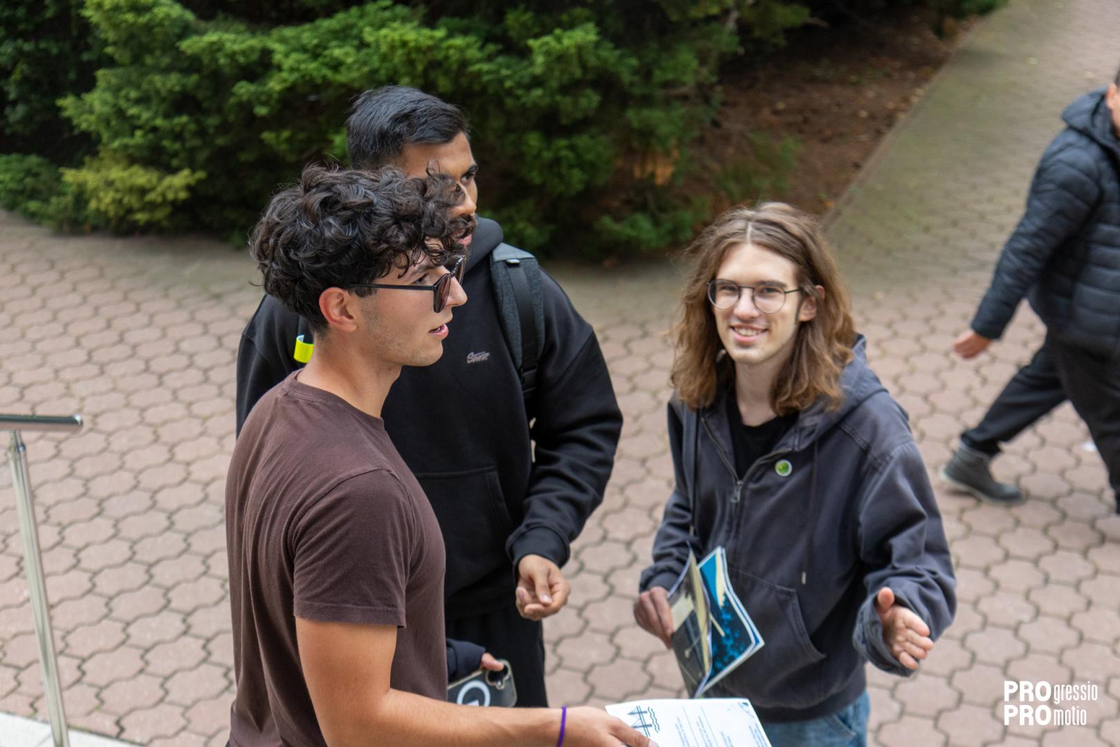Three students talk to each other on the sidewalk in front of bushes, holding campus game materials in their hands.