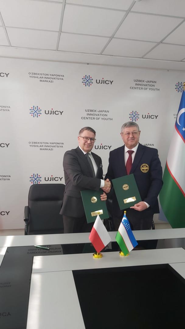 Dwaj mężczyźni stoją przy stole i podają sobie dłonie w geście porozumienia, trzymając zielone teczki ze złotym emblematem. Obaj patrzą w stronę obiektywu, a za nimi znajduje się ściana z wspomnianymi napisami i logo.