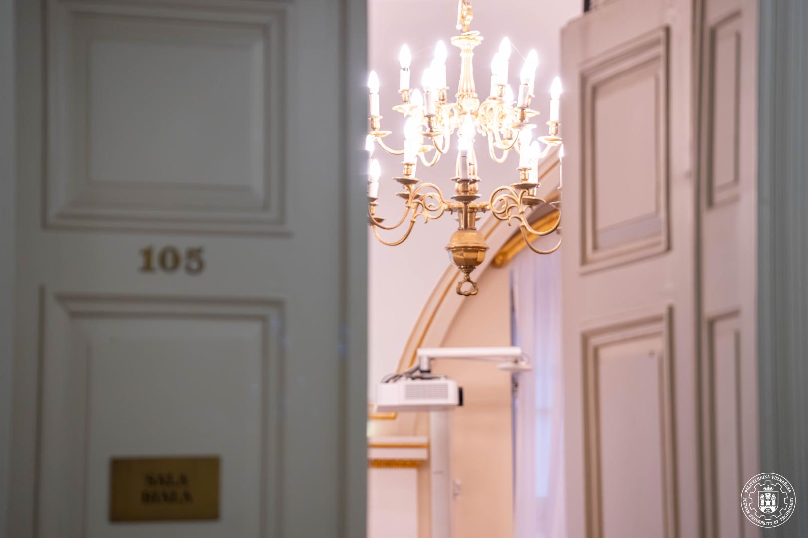 Widok uchylonych zdobionych białych drzwi sali z mosiężną tabliczką "SALA BIAŁA" i numerem 105, ukazujący wnętrze z żyrandolem, złoconymi zdobieniami i wiszącym projektorem na ścianie.
