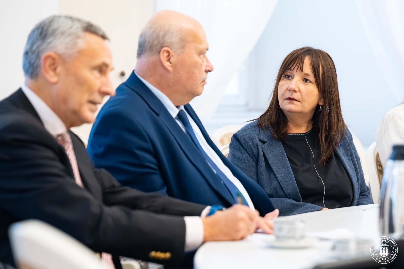 Trzy osoby siedzące przy okrągłym stole: po lewej starszy mężczyzna w różowym krawacie trzyma ręce na stole, w centrum mężczyzna w granatowym garniturze, a obok prof. Mistal w szaroniebieskiej marynarce spogląda na niego.