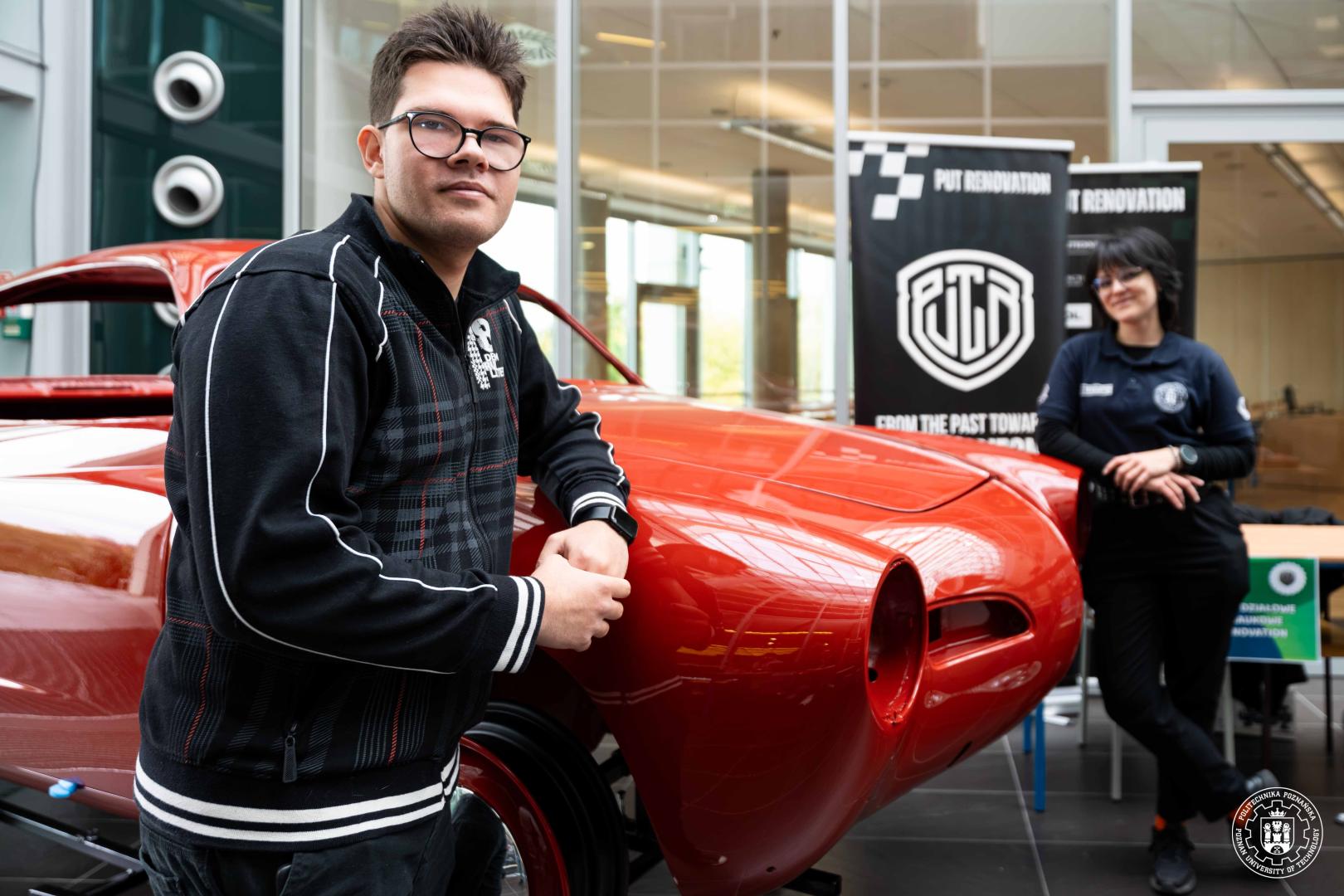Student opiera się o renowowaną karoserię czerwonego klasycznego samochodu. Baner z napisem: „PUT RENOVATION – FROM THE PAST TOWARDS THE FUTURE”.