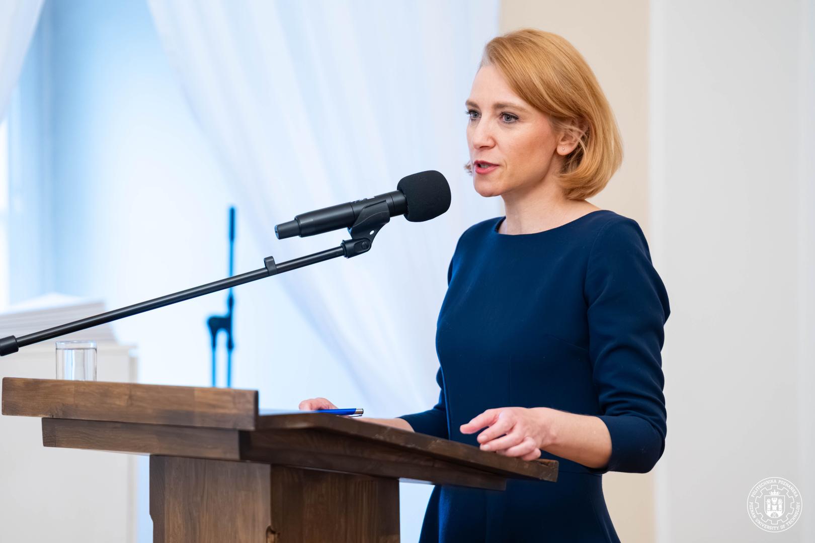 Kobieta w granatowej sukience przemawia za drewnianym pulpitem z mikrofonem, trzymając długopis, na tle jasnej ściany z firankami.