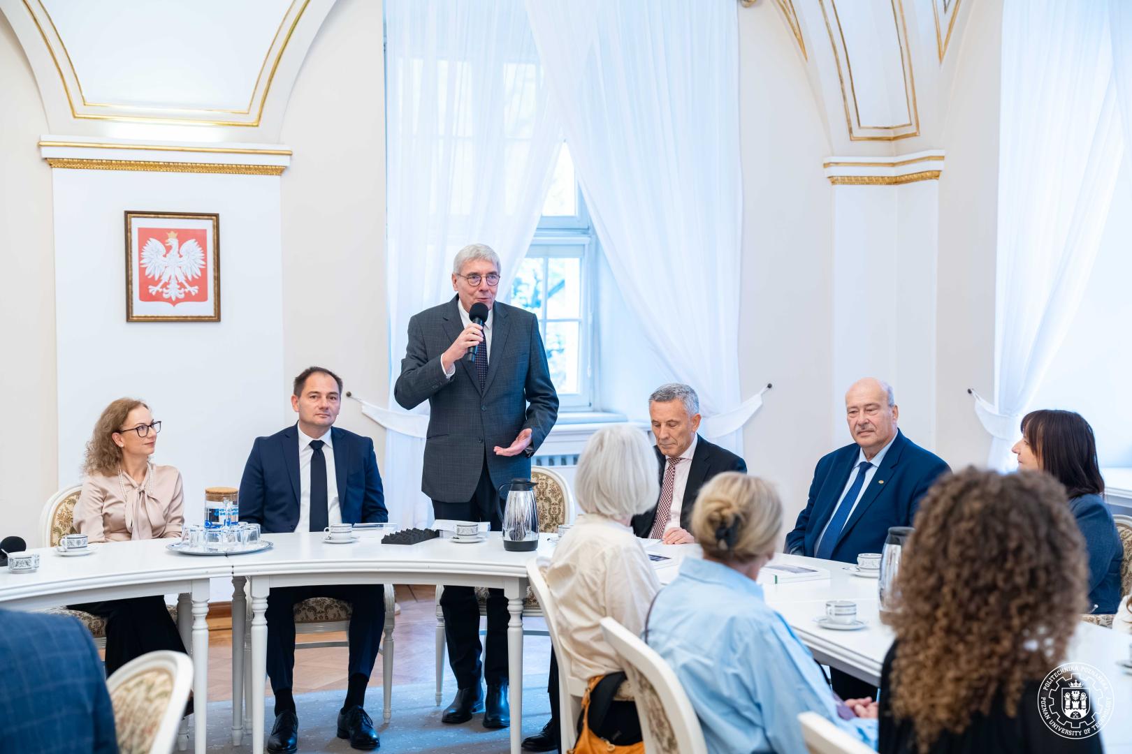 Mężczyzna w szarej marynarce stoi, trzyma mikrofon, przemawiając do grupy siedzących osób przy owalnym stole. W tle godło Polski w złotej ramie na ścianie.