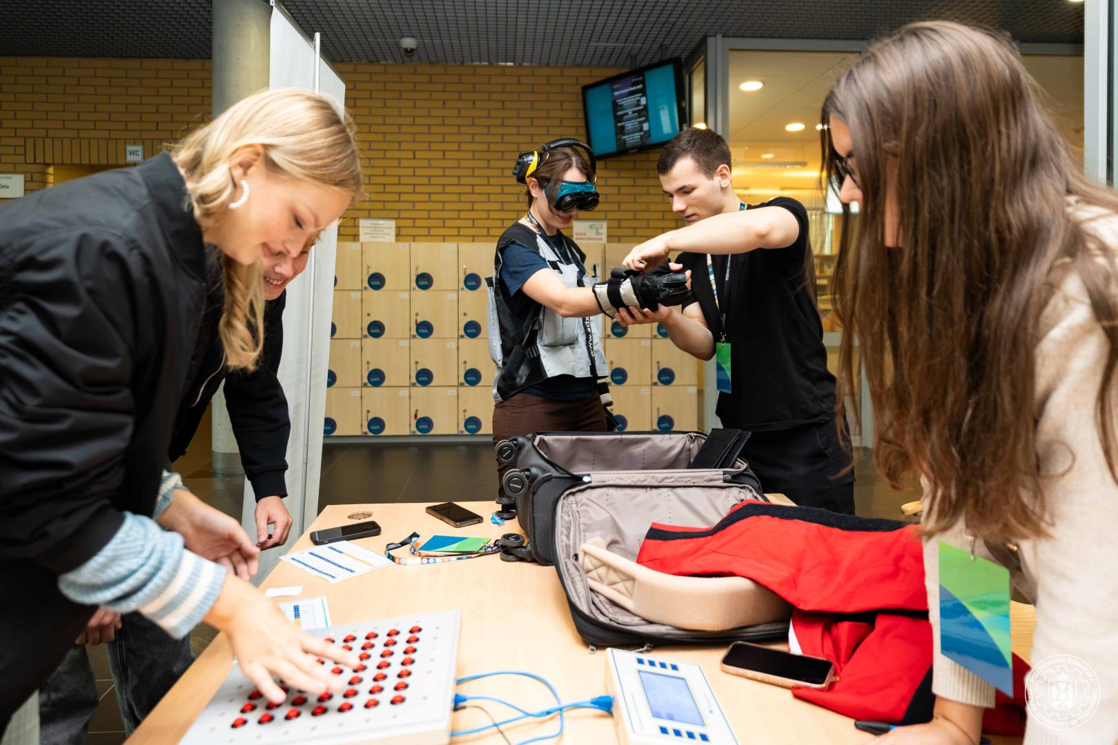 Studenci skupieni wokół stanowiska laboratoryjnego z panelem świecących czerwonych diod. Student obsługuje laptopa i dotyka panelu, a inna osoba robi zdjęcie telefonem.