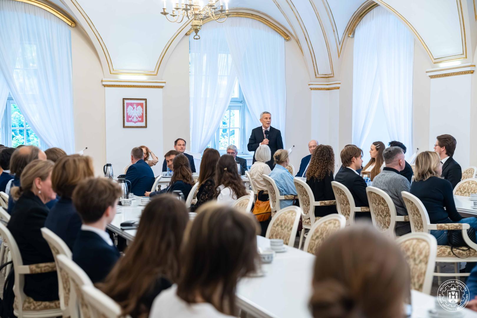 Mężczyzna w ciemnym garniturze przemawia do licznej widowni siedzącej przy białych stołach na zdobionych krzesłach - szeroki ujęcie