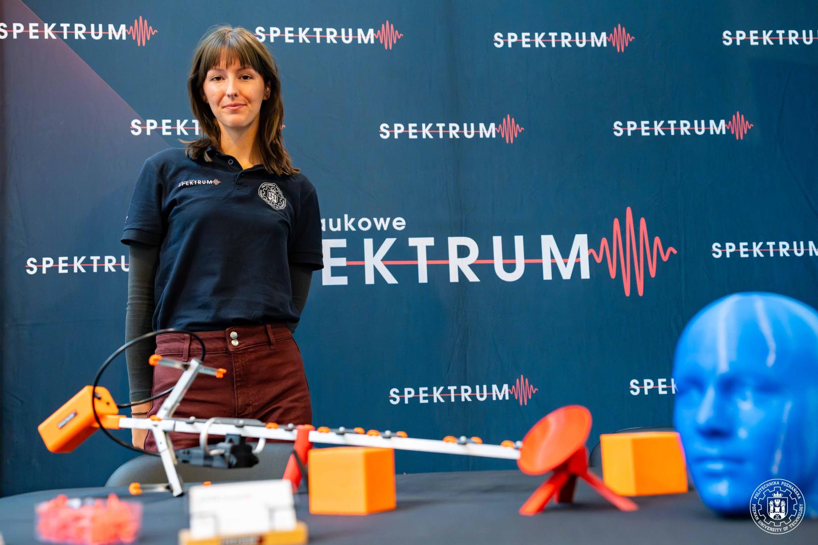 Kobieta w koszulce z logo "SPEKTRUM" stoi przed tłem z powtarzającym się napisem "SPEKTRUM", przed nią model techniczny z białych i pomarańczowych elementów.