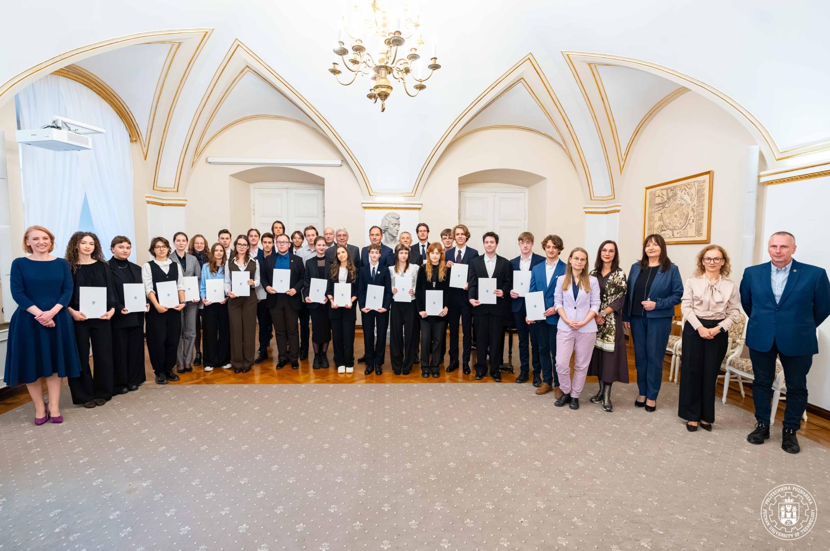 Kilkudziesięciu młodych uczestników oraz organizatorów ceremonii wręczenia stypendiów w reprezentacyjnej sali ze złoconym sklepieniem, trzymających białe teczki, ustawionych w kilku rzędach pod żyrandolem, pozują do grupowego zdjęcia.