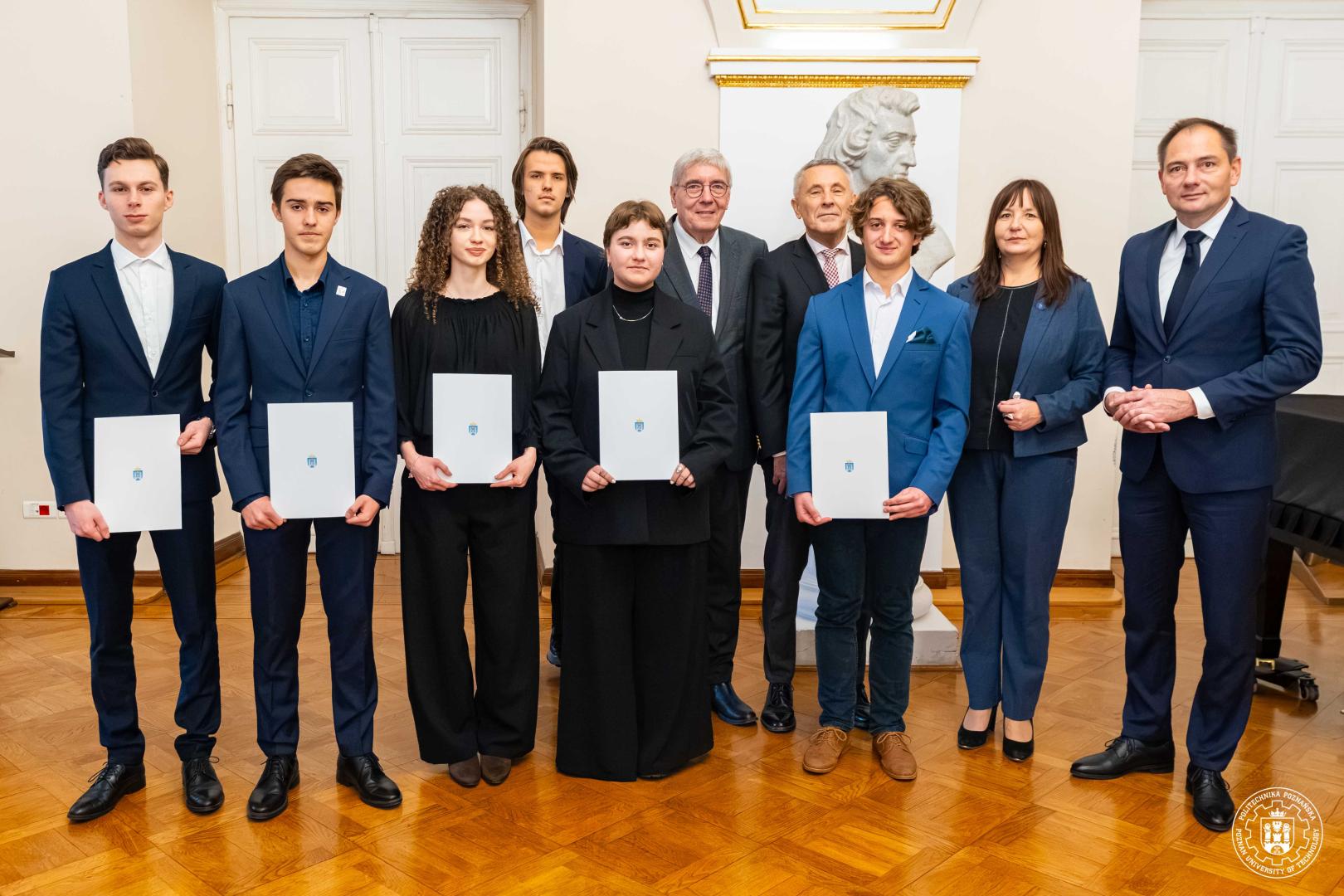 Grupa dziesięciorga osób w Sali Białej (wśród nich prof. Agnieszka Misztal), w tym sześcioro studentów w ciemnych strojach trzymających białe teczki z niebieskim logo, stojących w rzędzie przed popiersiem na cokole i fortepianem widocznym w tle.