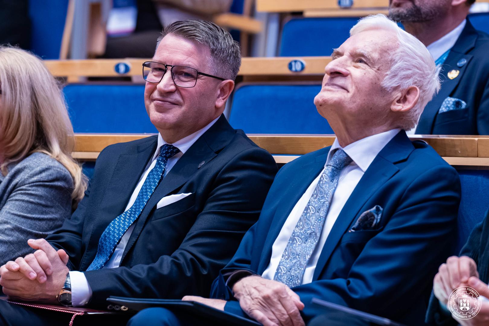 Rektor PP i prof. Buzek siedzą w pierwszym rzędzie sali konferencyjnej w eleganckich garniturach, trzymają teczki na kolanach.