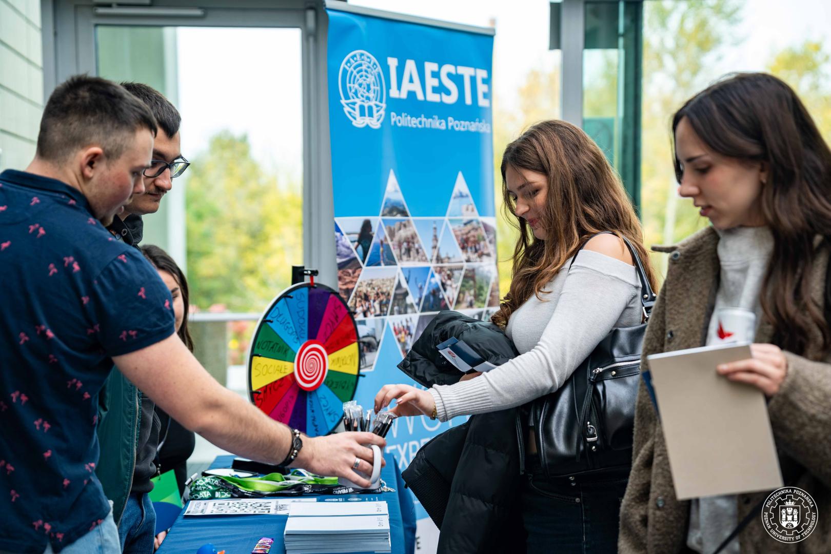 Studenci przy niebieskim stoisku organizacji IAESTE z kołem fortuny, na którym leżą gadżety promocyjne.