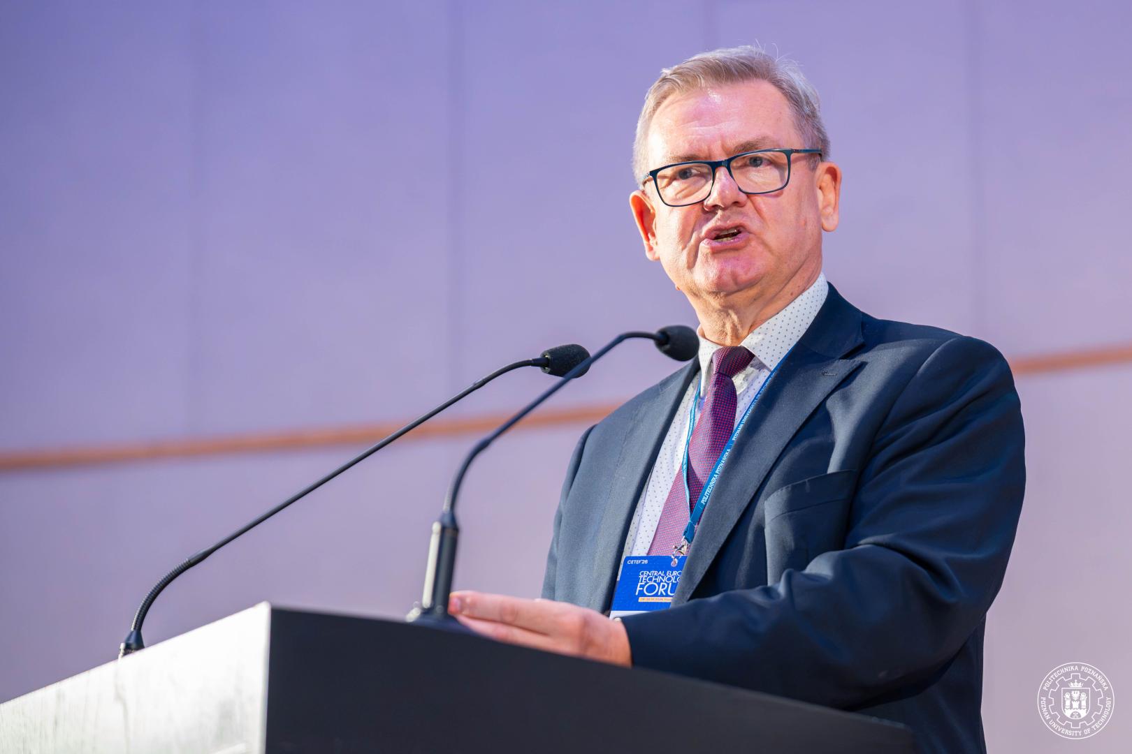 Prezes IZ TECH w ciemnym garniturze i czerwonym krawacie przemawia za mównicą. W tle widoczna jest jasna, gładka ściana auli z delikatnymi pionowymi pasami w odcieniu szarości.