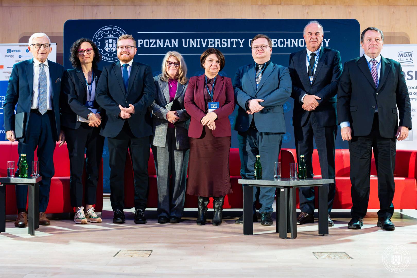 Ośmiu panelistów pozuje w rzędzie na scenie do grupowego zdjęcia przed banerem z napisem "POZNAN UNIVERSITY OF TECHNOLOGY", obok nich niskie stoliki z butelkami wody.