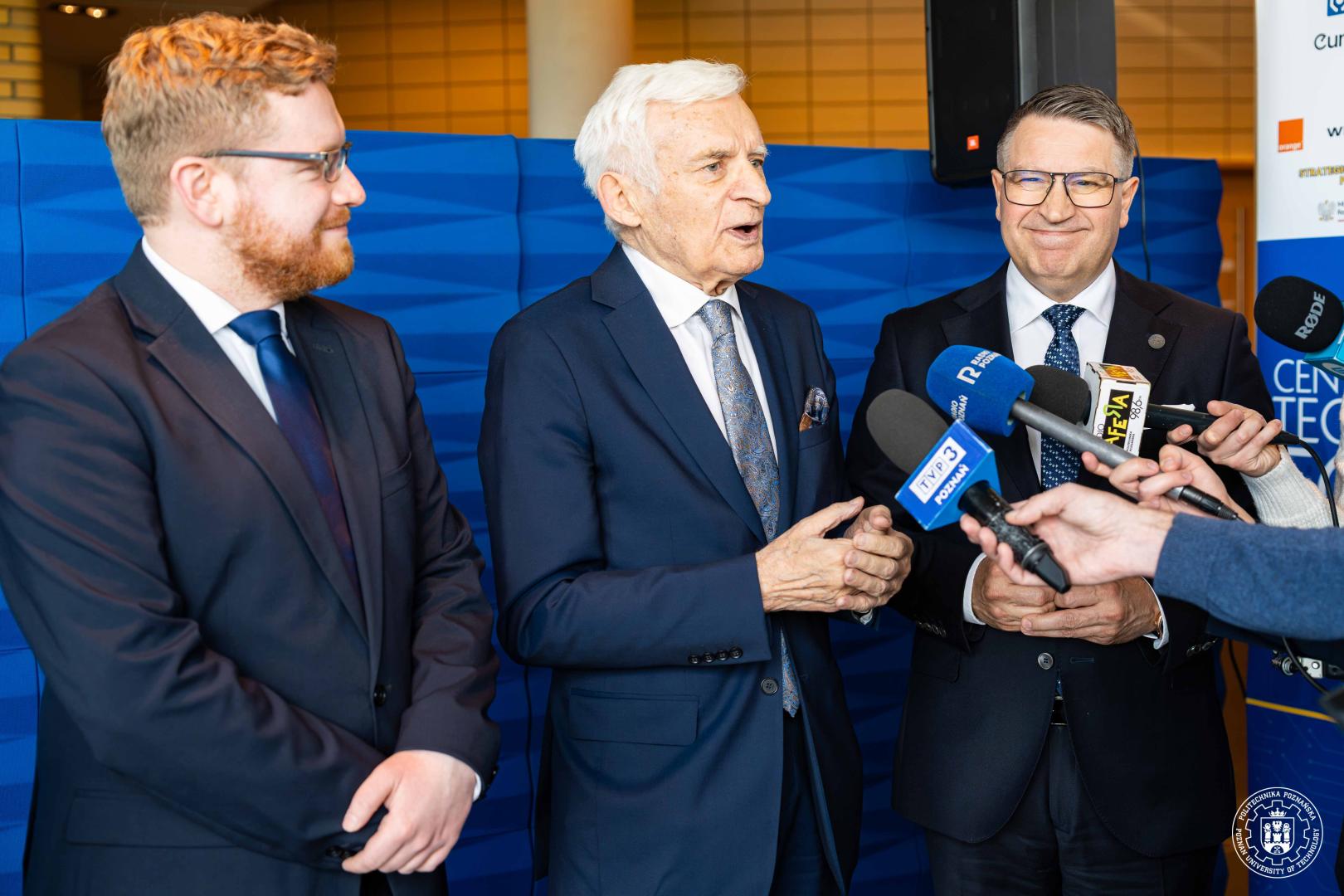 Rektor PP, prof. Jerzy Buzek i Dariusz Standerski stoją w otoczeniu dziennikarzy: starszy mężczyzna z siwymi włosami przemawia i gestykuluje, obok mikrofony z logo m.in. "TVP3 Poznań" na tle niebieskiej materiałowej ścianki.