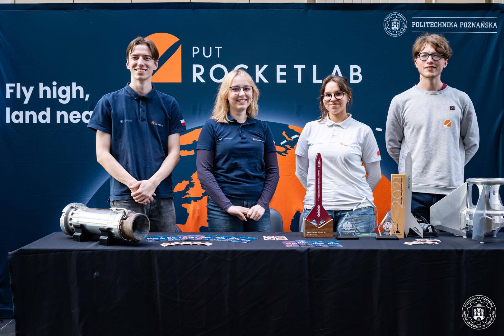 Czterej studenci stoją przed stołem z modelem rakiety i trofeami, wszyscy w koszulkach z logo "PUT Rocketlab", w tle baner z hasłem "Fly high, land near".
