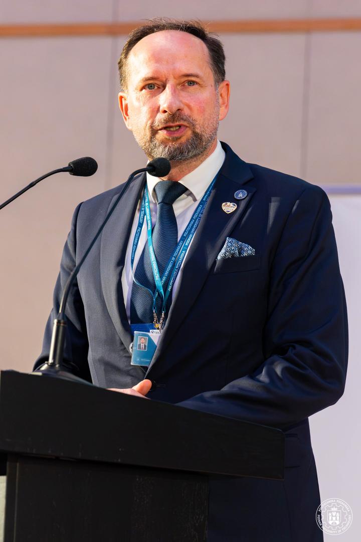 Prof. Arkadiusz Ptak z krótką brodą i identyfikatorem na szyi przemawia za pulpitem z mikrofonem, na klapie marynarki widoczna metalowa odznaka z logo PP.
