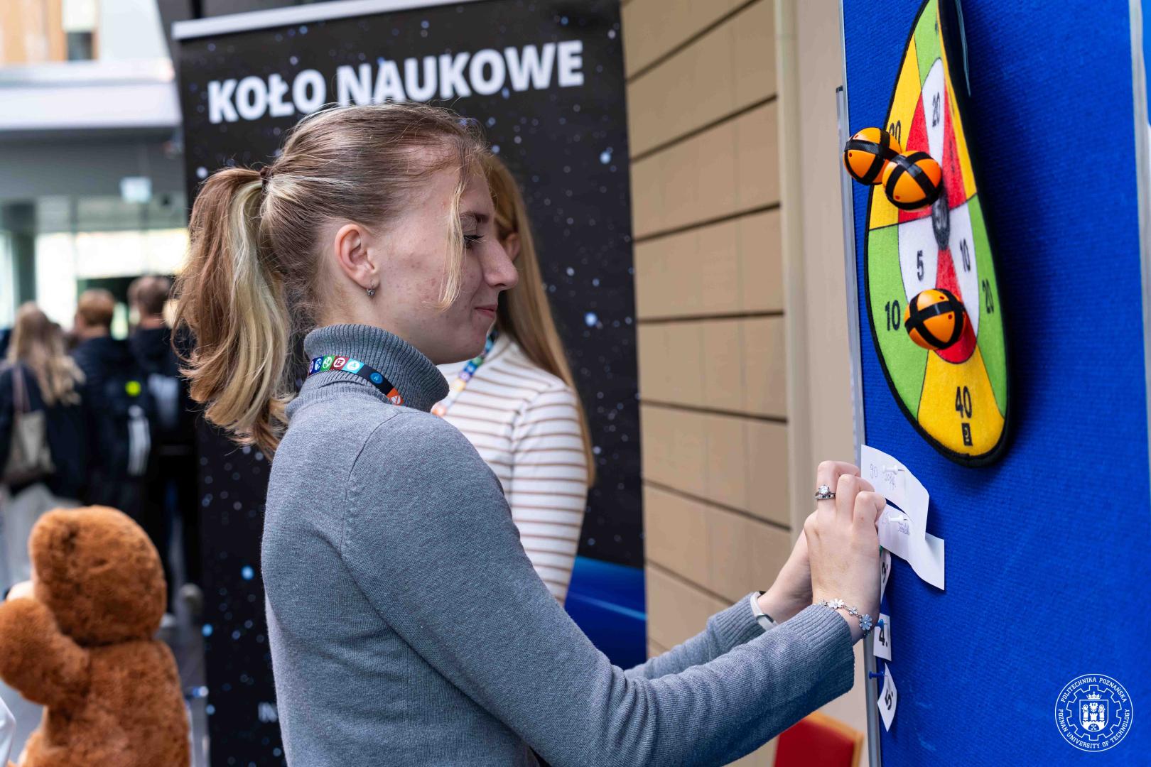 Młoda kobieta przypina kartkę do niebieskiej tablicy przy rzutkach na rzepy na stoisku koła naukowego.