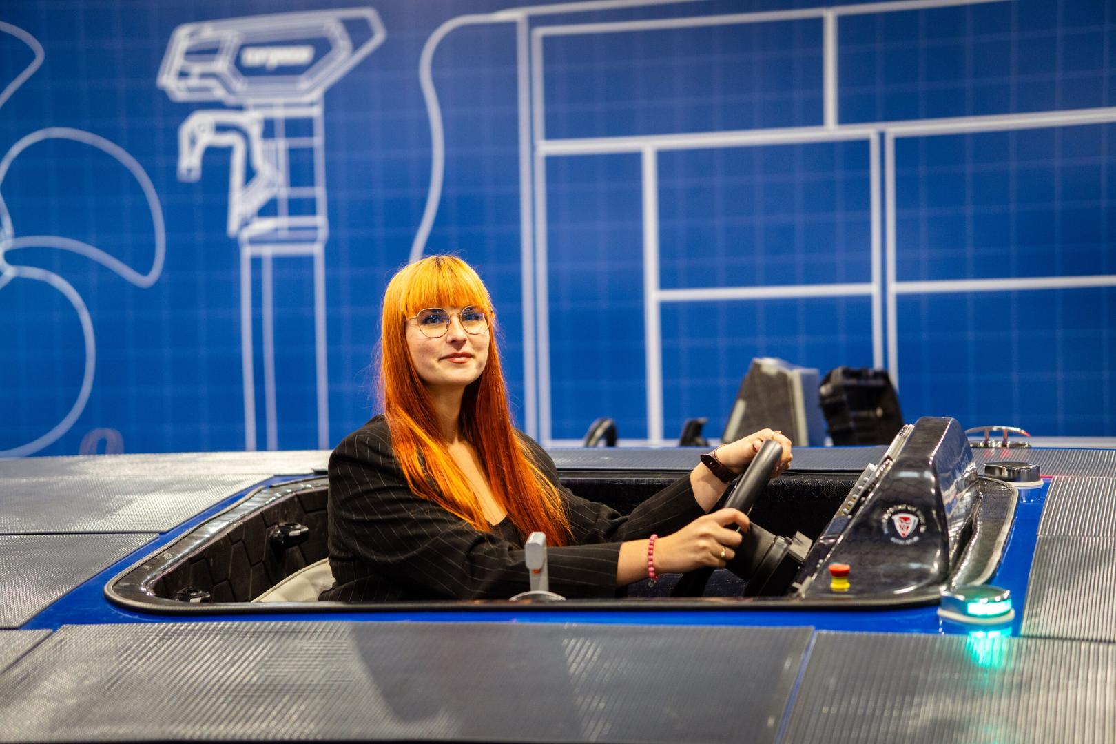 Kobieta w ciemnej marynarce siedzi w łodzi PUT Powertrain, trzymając koło sterowe, na tle niebieskiej ściany stoiska z białymi schematami technicznymi.