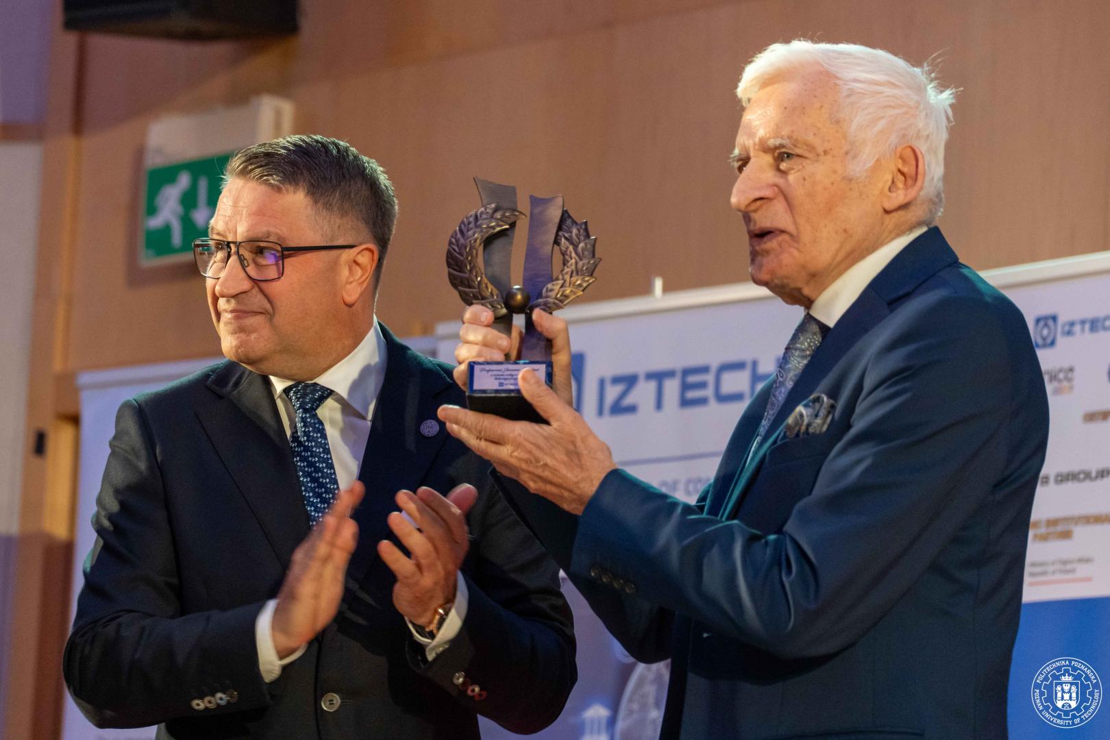 Rektor PP i prof. J. Buzek stoi obok siebie: jeden klaszcze, drugi trzyma statuetkę w kształcie liści laurowych na czarnej podstawie, w tle baner z logo "IZTECH".