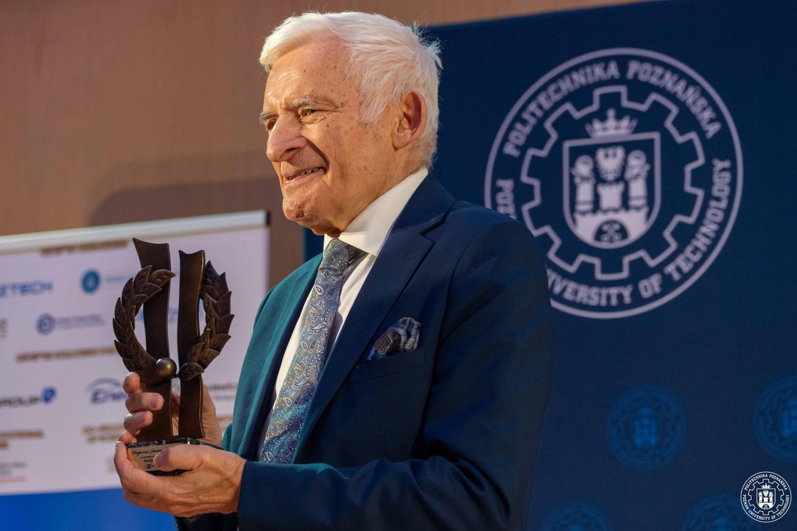 Proj. Jerzy Buzek w ciemnoniebieskim garniturze trzyma metalową, brązowawą statuetkę w kształcie liści laurowych na scenie z logo Politechniki Poznańskiej w tle.