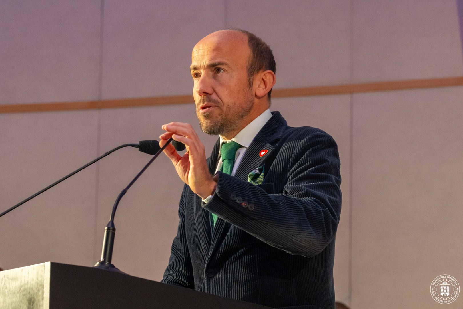 Borys Budka w ciemnej, sztruksowej marynarce z zielonym krawatem przemawia za mównicą, trzymając mikrofon. Na klapie marynarki widoczna jest przypinka w kształcie serca.
