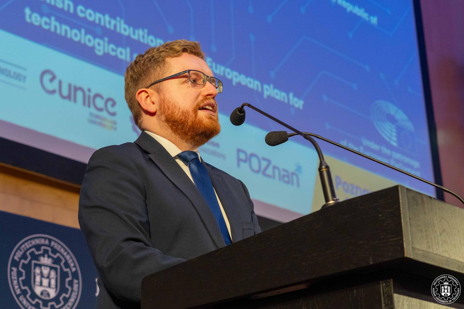 Dariusz Standerski z rudą brodą i okularami przemawia za pulpitem z mikrofonami, na ekranie w tle widoczne fragmenty prezentacji z napisami i logo partnerów konferencji.
