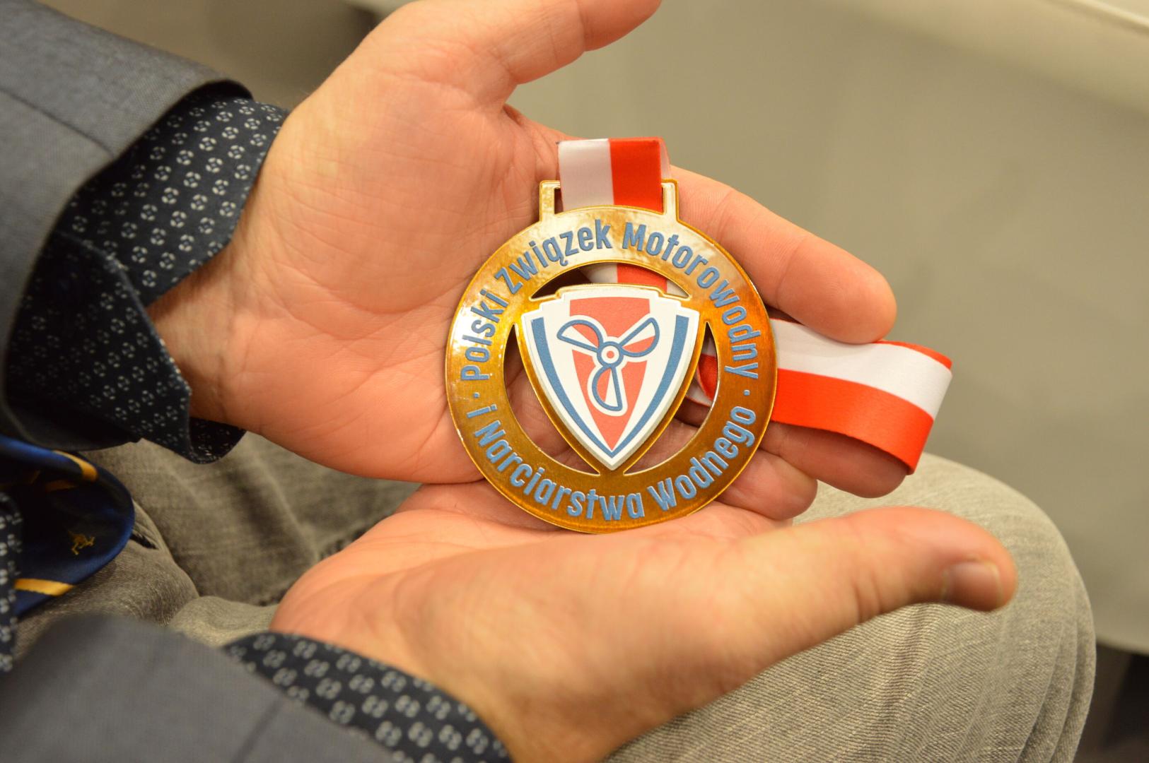 Bliskie ujęcie na dłonie pokazujące do aparatu złoty medal z napisem "POLSKI ZWIĄZEK MOTOROWODNY i NARCIARSTWA WODNEGO" na biało-czerwonej wstążce, z symbolem śmigła na tarczy.