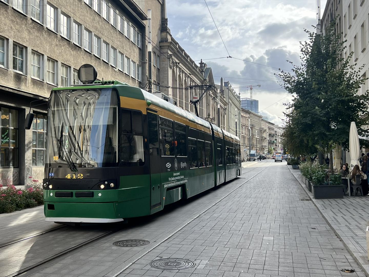 Zielono-żółty tramwaj MPK Poznań jadący ulicą miasta, z widocznymi na boku logo partnerów projektu oraz informacją o badaniach oświetlenia.