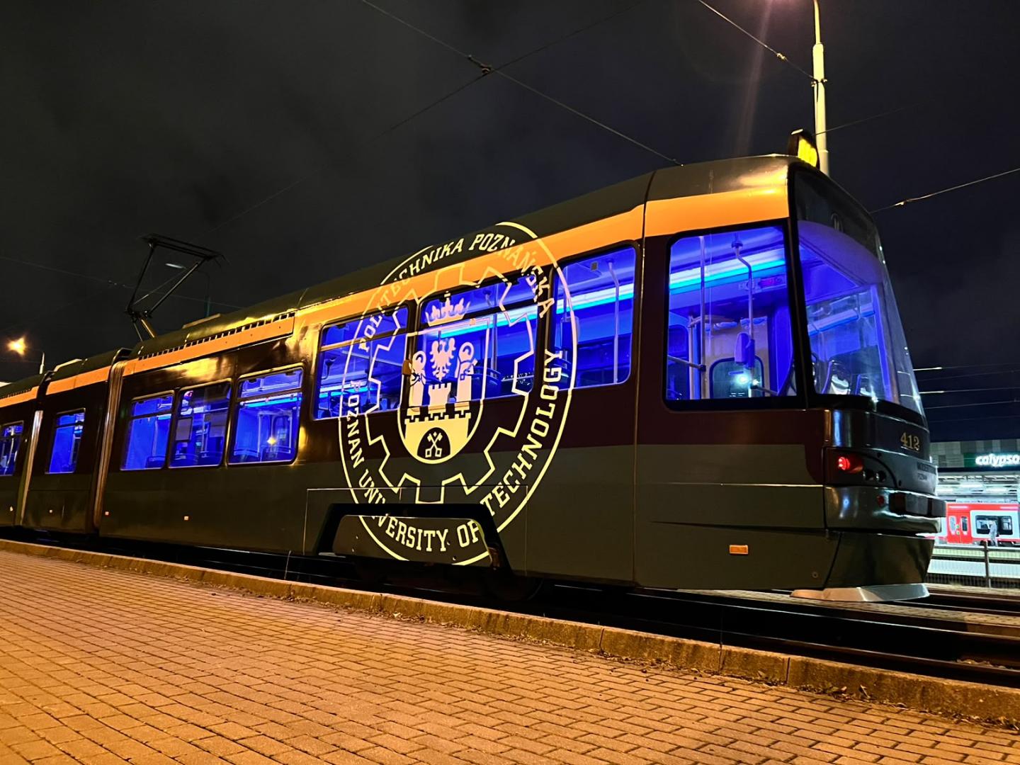 Tramwaj MPK Poznań w nocy z podświetlonym na niebiesko wnętrzem oraz dużym logo Politechniki Poznańskiej na boku pojazdu.