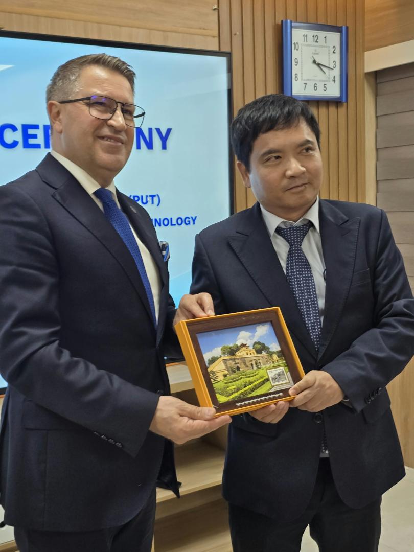 Rektor Politechniki Poznańskiej oraz Rektor PTIT trzymają oprawiony w ramkę obrazek przedstawiający zabytkowy obiekt, stojąc na tle ekranu z napisami.