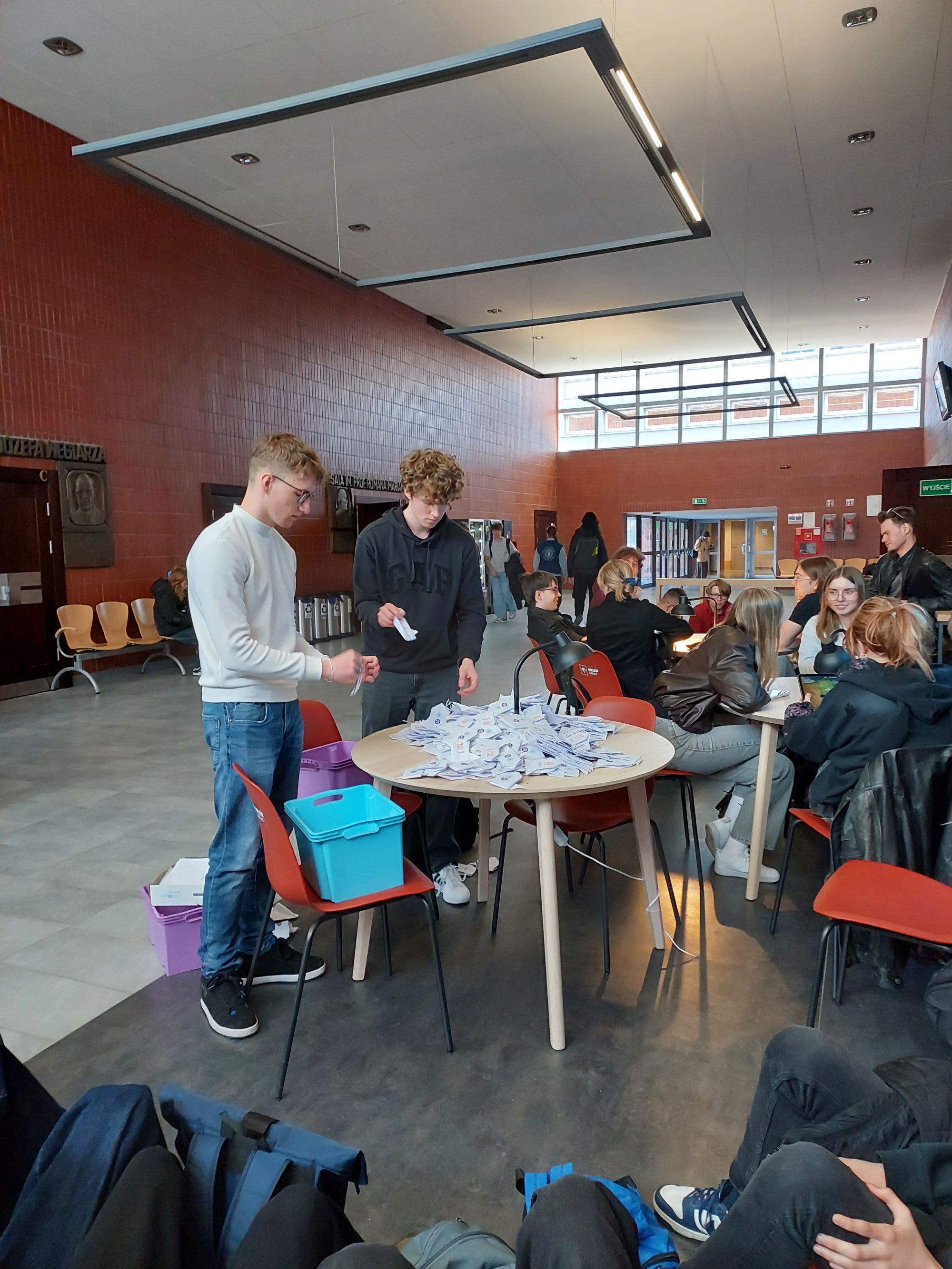 Grupa studentów w przestronnym holu akademickim składa białe papierowe rakiety origami przy okrągłym stole podczas bicia Rekordu Polski.