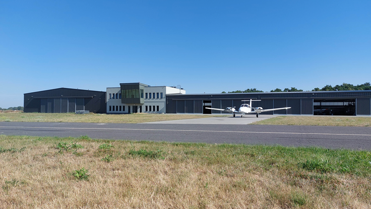 Dwa hangary połąoczone budynkiem zaplecza badawczego. Przed hangarem z prawej stoi mały biały samolot.
