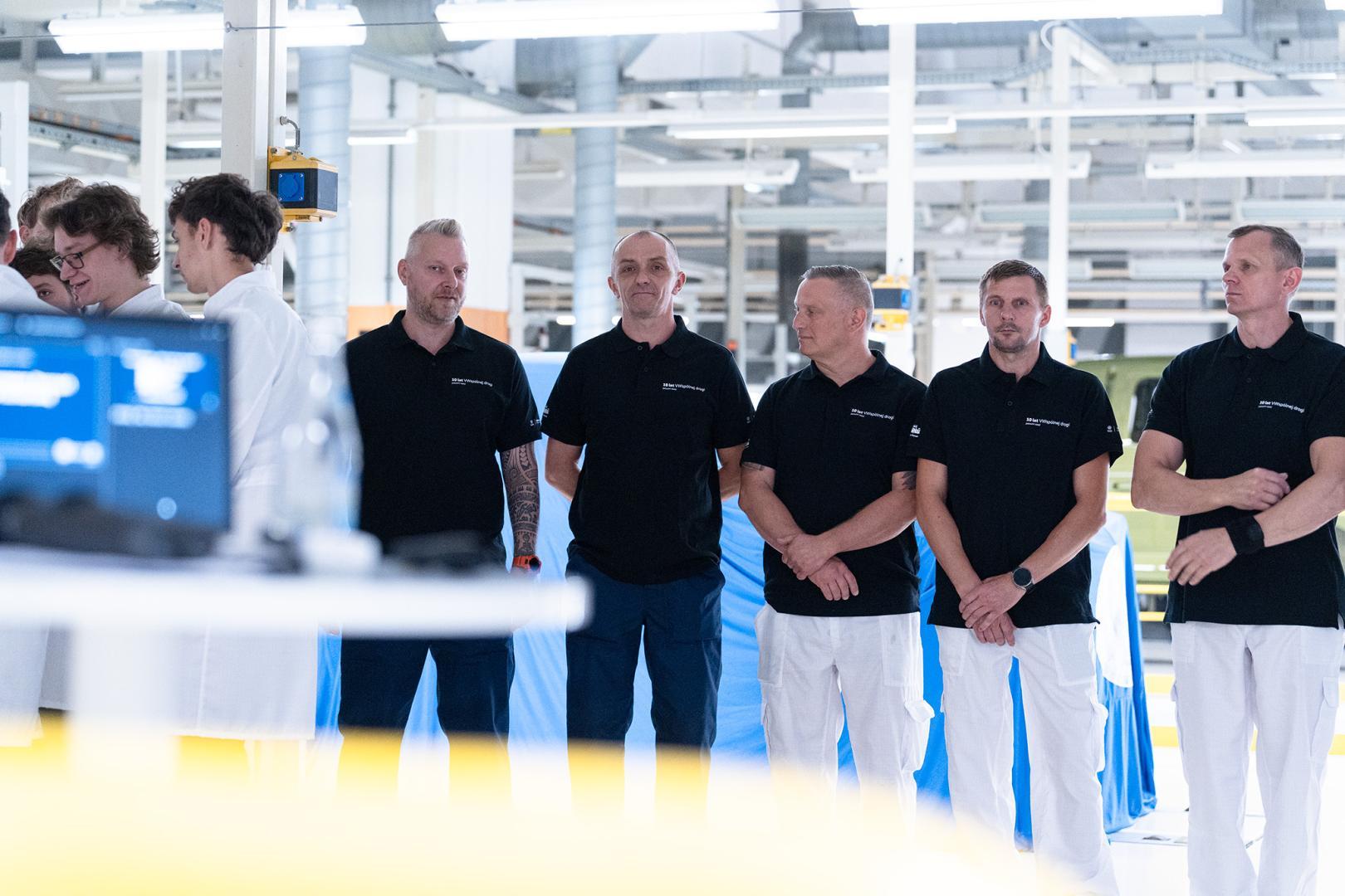 Pracownicy lakierni Volkswagen Poznań w czarnych koszulkach polo stoją w rzędzie na tle przykrytej niebieską płachtą karoserii samochodu.