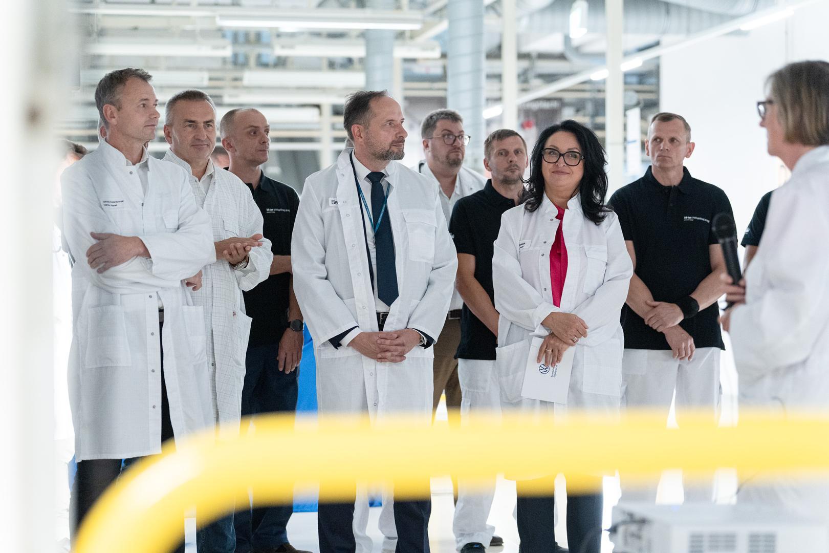 Zarząd Volkswagen Poznań oraz władze Politechniki Poznańskiej w białych fartuchach słuchają przemówienia podczas uroczystości w lakierni.