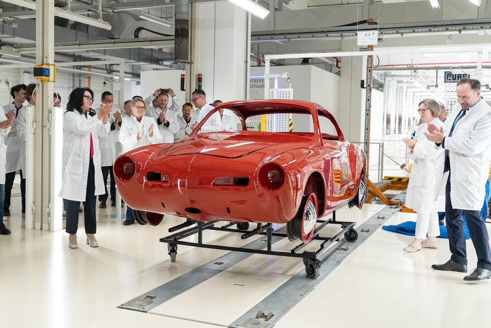 Odsłonięta, błyszcząca, czerwona karoseria zabytkowego Volkswagena Karmann Ghia, stojąca na wózku technologicznym w lakierni pośród klaszczących uczestników uroczystości.