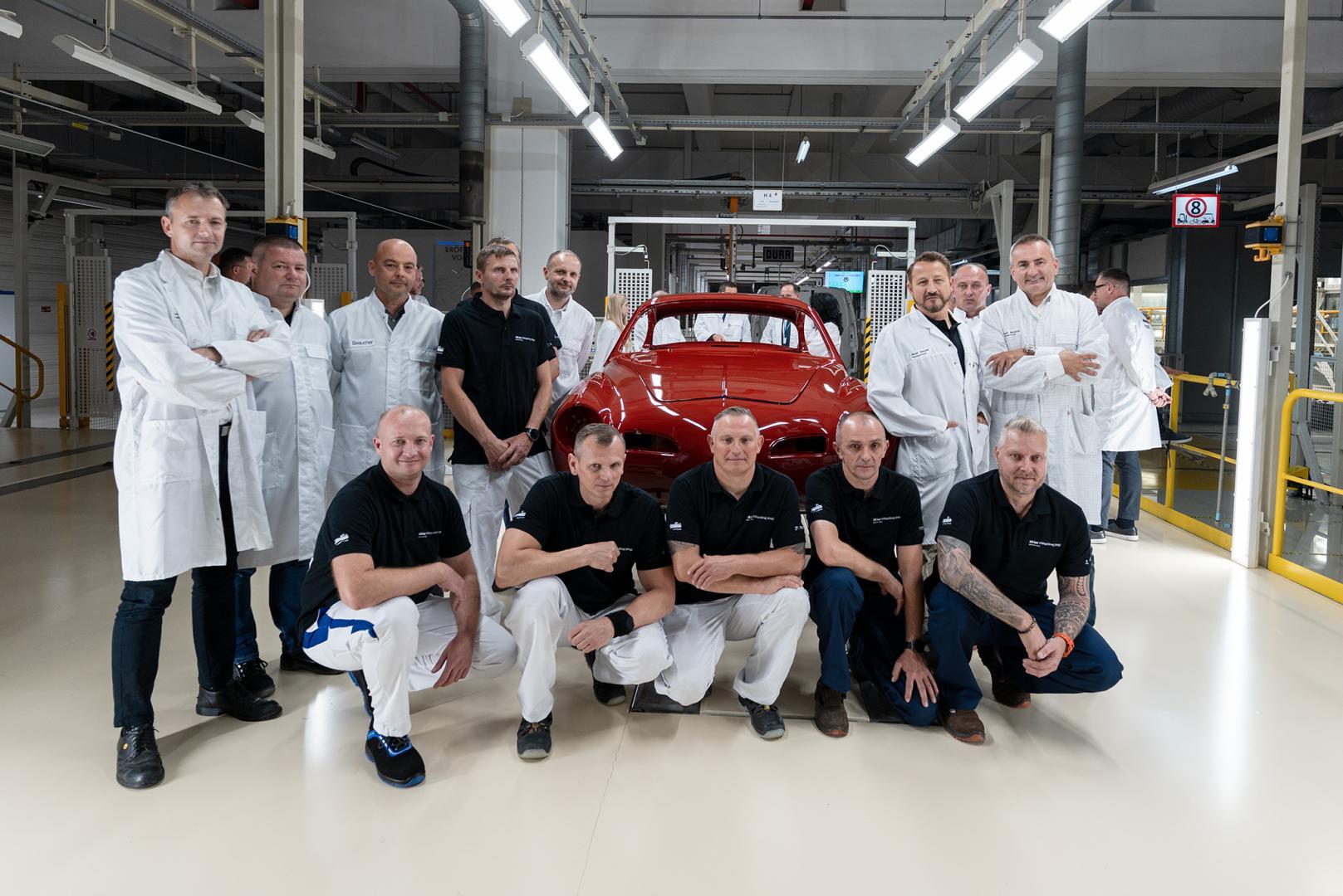 Pracownicy fabryki pozują wokół odrestaurowanego czerwonego Volkswagena Karmann Ghia w hali produkcyjnej.