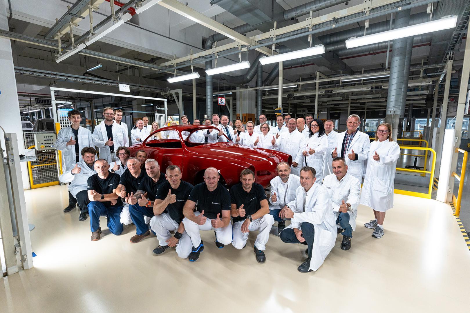Pracownicy fabryki pozują wokół odrestaurowanego czerwonego Volkswagena Karmann Ghia w hali produkcyjnej.