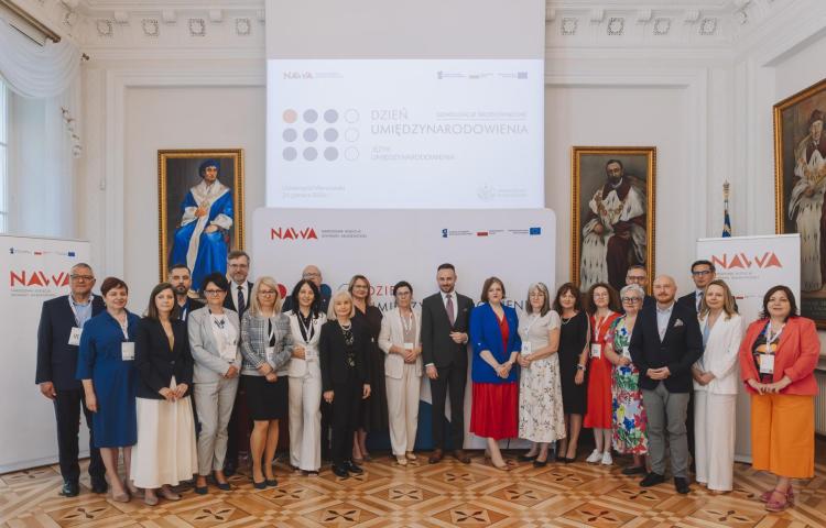 Grupowe zdjęcie uczestników wydarzenia NAWA „Dzień Umiędzynarodowienia” na tle sceny i materiałów konferencyjnych.
