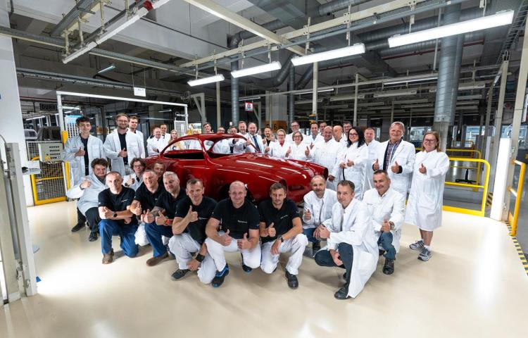 Pracownicy fabryki pozują wokół odrestaurowanego czerwonego Volkswagena Karmann Ghia w hali produkcyjnej.