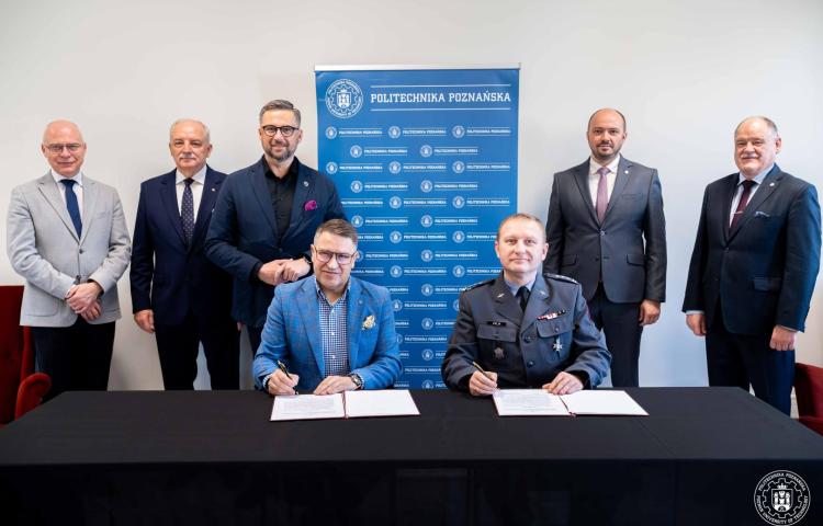 Dwaj mężczyźni siedzą przy stole i składają podpisy na dokumentach, a za nimi stoi w rzędzie pięciu mężczyzn (przedstawiciele uczelni i ministerstw) na tle niebieskiego baneru z logo uczelni - wszyscy pozują do zdjęcia.
