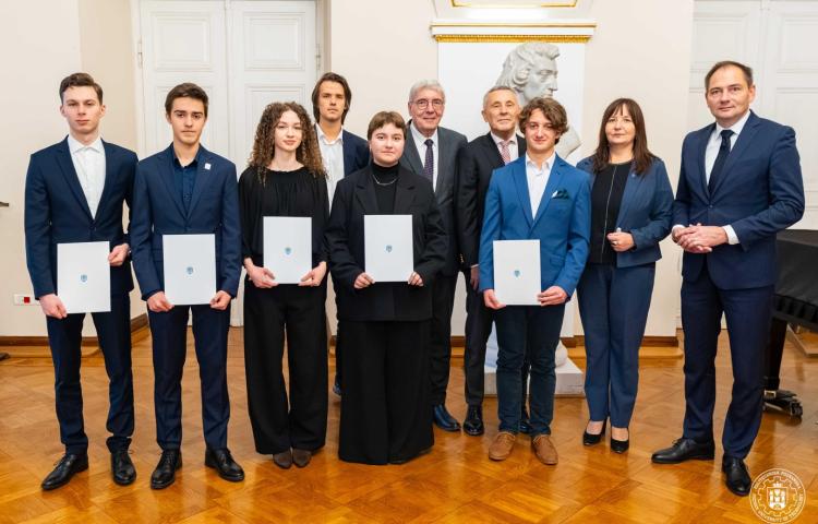 Grupa dziesięciorga osób w Sali Białej (wśród nich prof. Agnieszka Misztal), w tym sześcioro studentów w ciemnych strojach trzymających białe teczki z niebieskim logo, stojących w rzędzie przed popiersiem na cokole i fortepianem widocznym w tle.